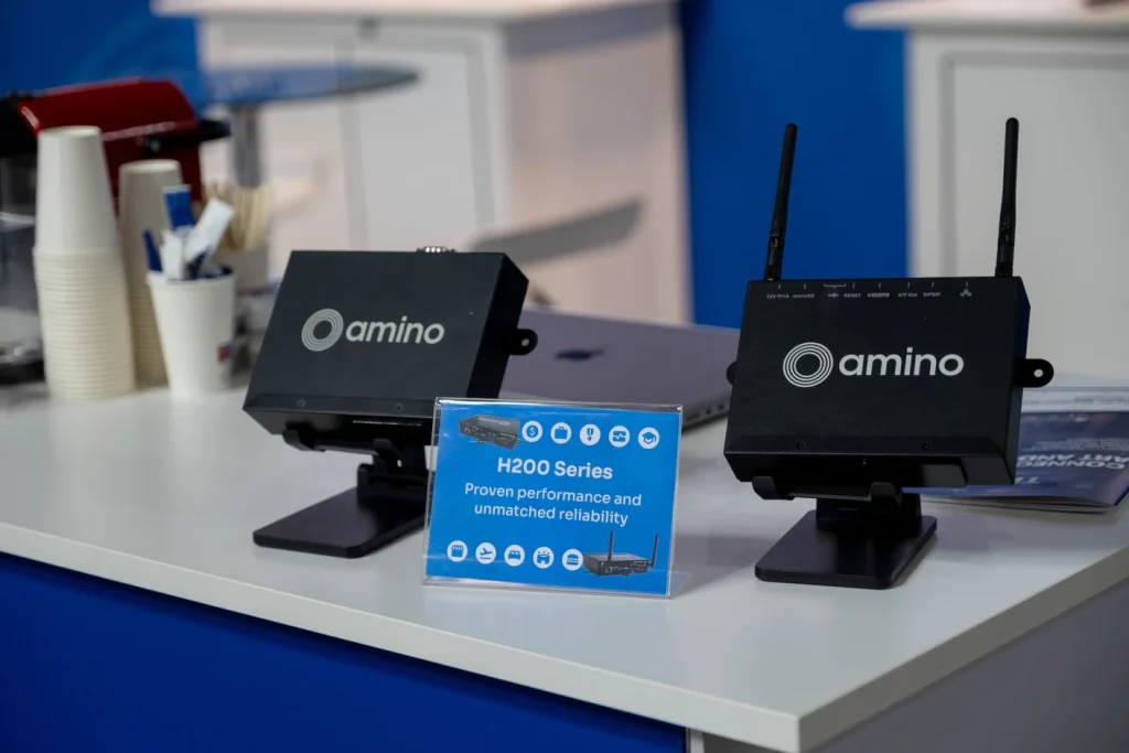 Two black Amino H200 Series devices with antennas are displayed on a table beside a blue information sign at an exhibition booth.