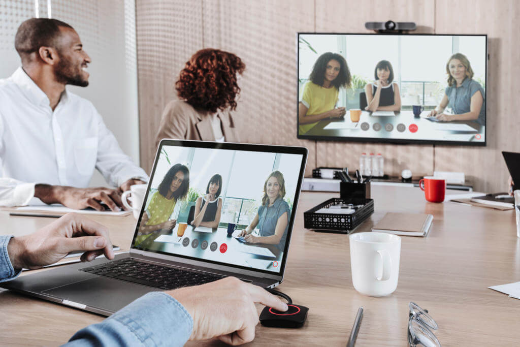 A group of people in an office participates in a video conference call with three colleagues displayed on a laptop and large screen.
