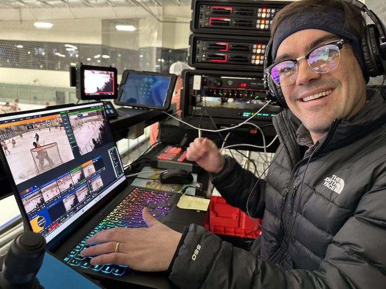 A person wearing headphones and glasses operates a laptop, NETGEAR M4250 AV Switches, and audio-visual equipment at a hockey rink, with multiple screens by StreamGeeks displaying live ice-hockey coverage.