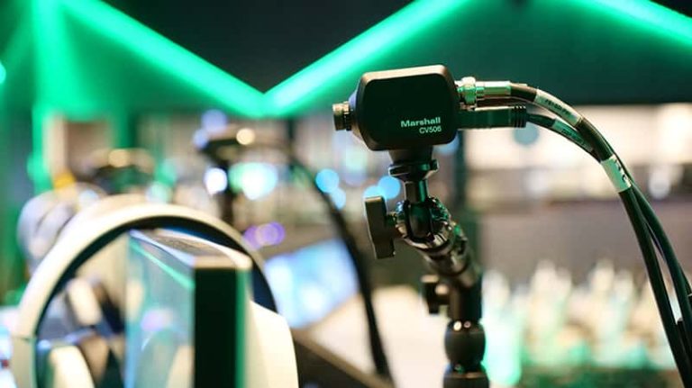 Close-up of a Marshall CV506 camera mounted on a flexible arm in an immersive AV space, with green LED lighting and monitors glowing in the background.