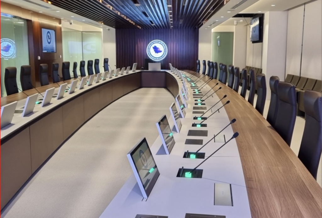A modern, empty conference room in a corporate headquarters features a long oval table with many chairs, microphones, and monitors. A large emblem is displayed on the wall at the end of the room, showcasing a recent boardroom upgrade.