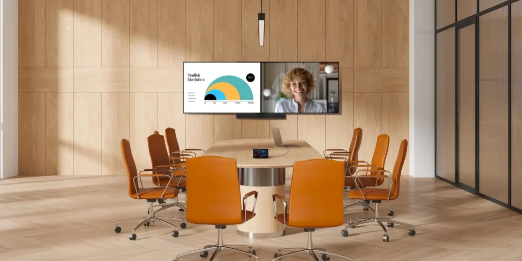 A modern conference room with eight orange chairs, a wooden table, and a large screen displaying a video call and a graph.