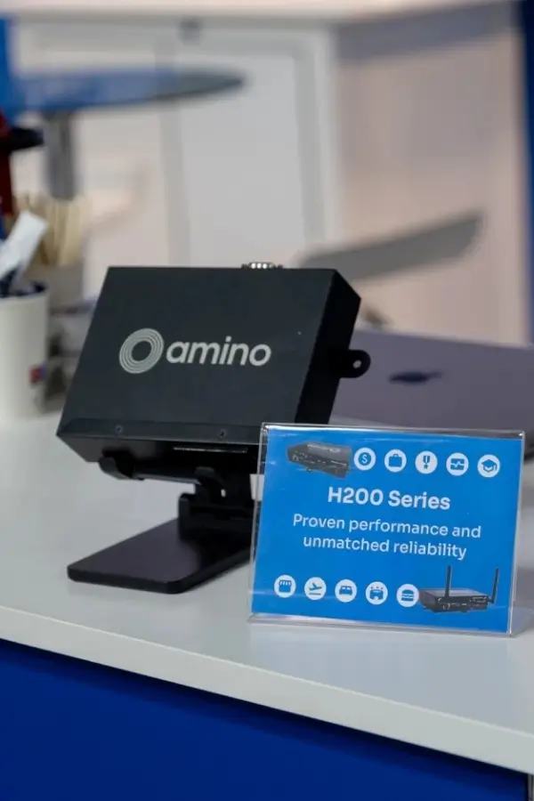 A black Amino device is displayed on a stand next to a sign reading "H200 Series—Proven performance and unmatched reliability" on a blue counter.