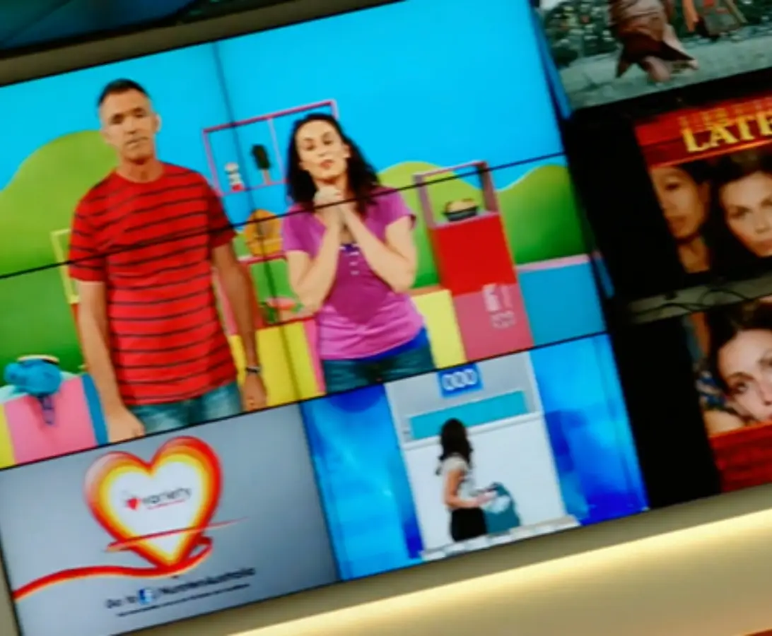 A wall of television screens displays various programs, including a man and woman on a colorful set, a heart logo, and scenes from other shows.