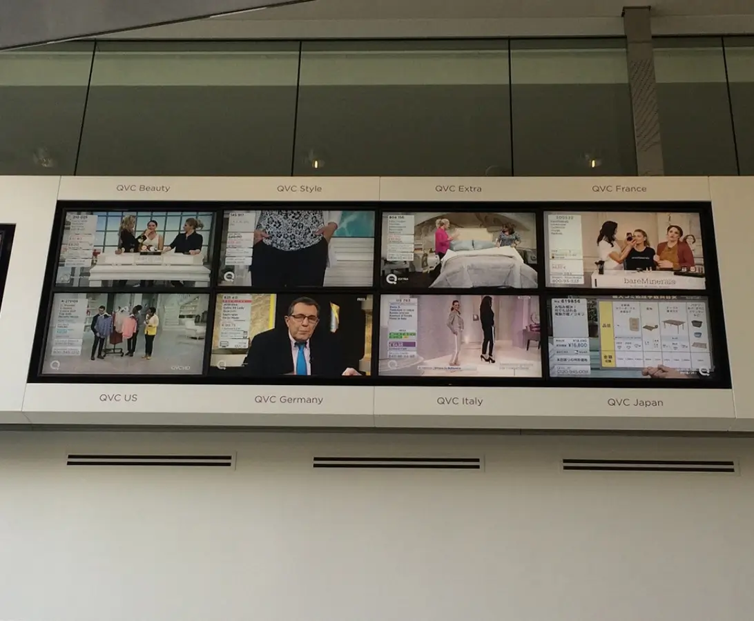 A wall-mounted display shows six TV screens tuned to different QVC shopping channels, labeled by region: US, Germany, Italy, Japan, Style, Extra, Beauty, and France.
