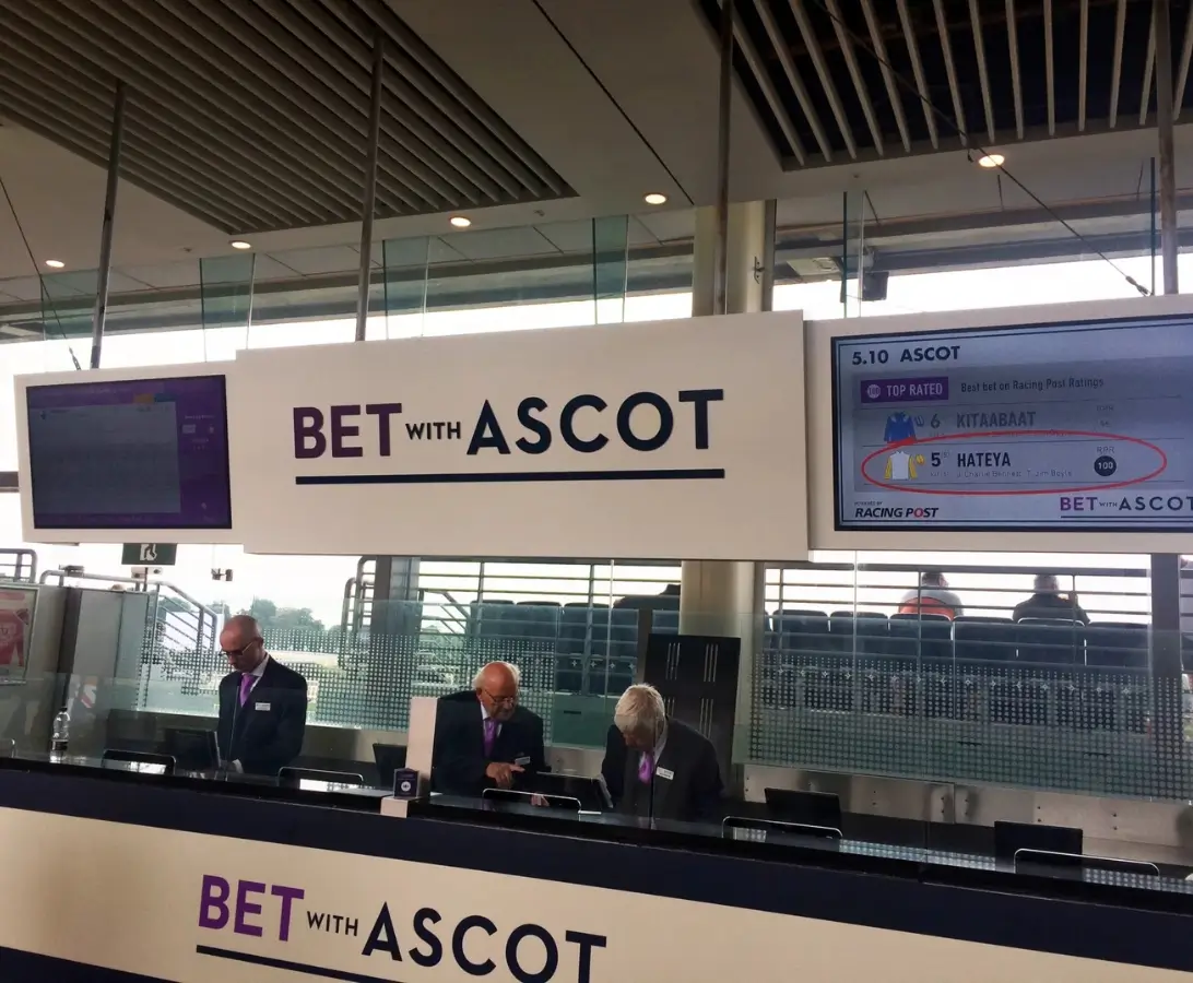 Three men sit behind a counter at a racetrack betting area with "BET WITH ASCOT" signage and a screen displaying race information.