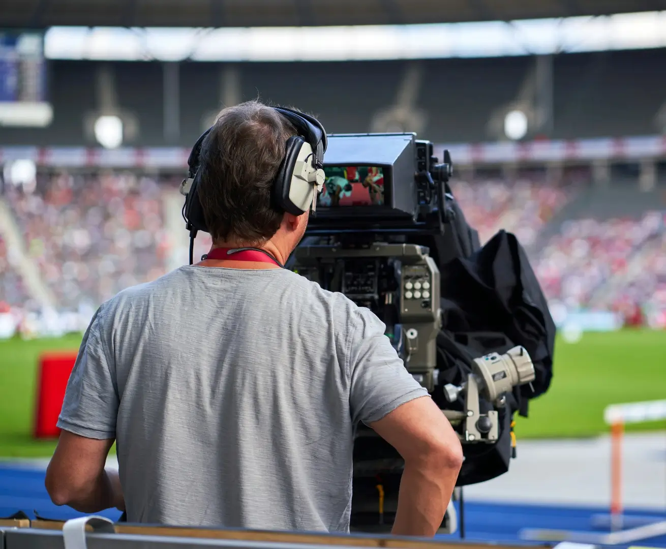 A camera operator wearing headphones films a sporting event in a stadium, focusing the camera on the field with blurred spectators in the background.