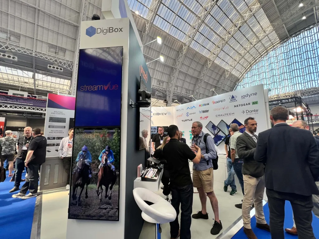 People interact at a busy tech expo booth with DigiBox branding, digital displays, and informational materials in a large, well-lit exhibition hall.