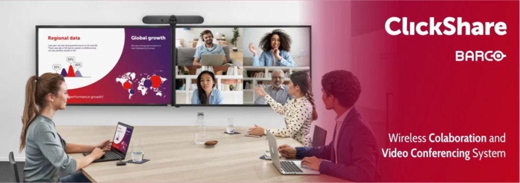 Four people in a conference room using laptops and a wireless presentation system, with two large screens displaying a data presentation and a video call. The screen shows the text "ClickShare" and "Barco.