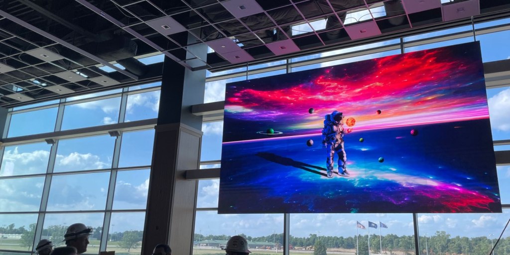 A large indoor screen displays a colorful, cosmic-themed image featuring an astronaut and planets. The setting has a high ceiling, large windows, and a few people wearing hard hats visible.