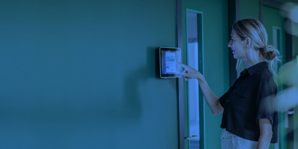 A person in a black shirt and beige pants uses a wall-mounted touchscreen device in a hallway with green walls and doors.