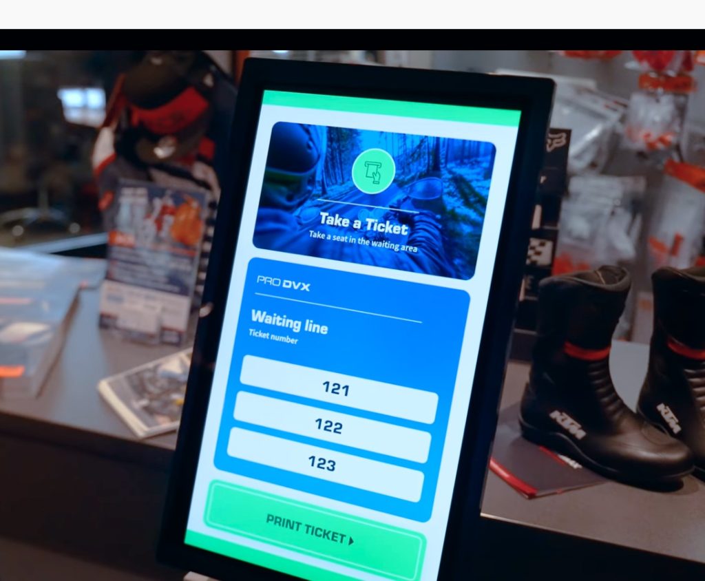 A digital kiosk screen displays a "Take a Ticket" interface with a waiting line, showing ticket numbers 121, 122, and 123. Nearby sports equipment is visible in the background.
