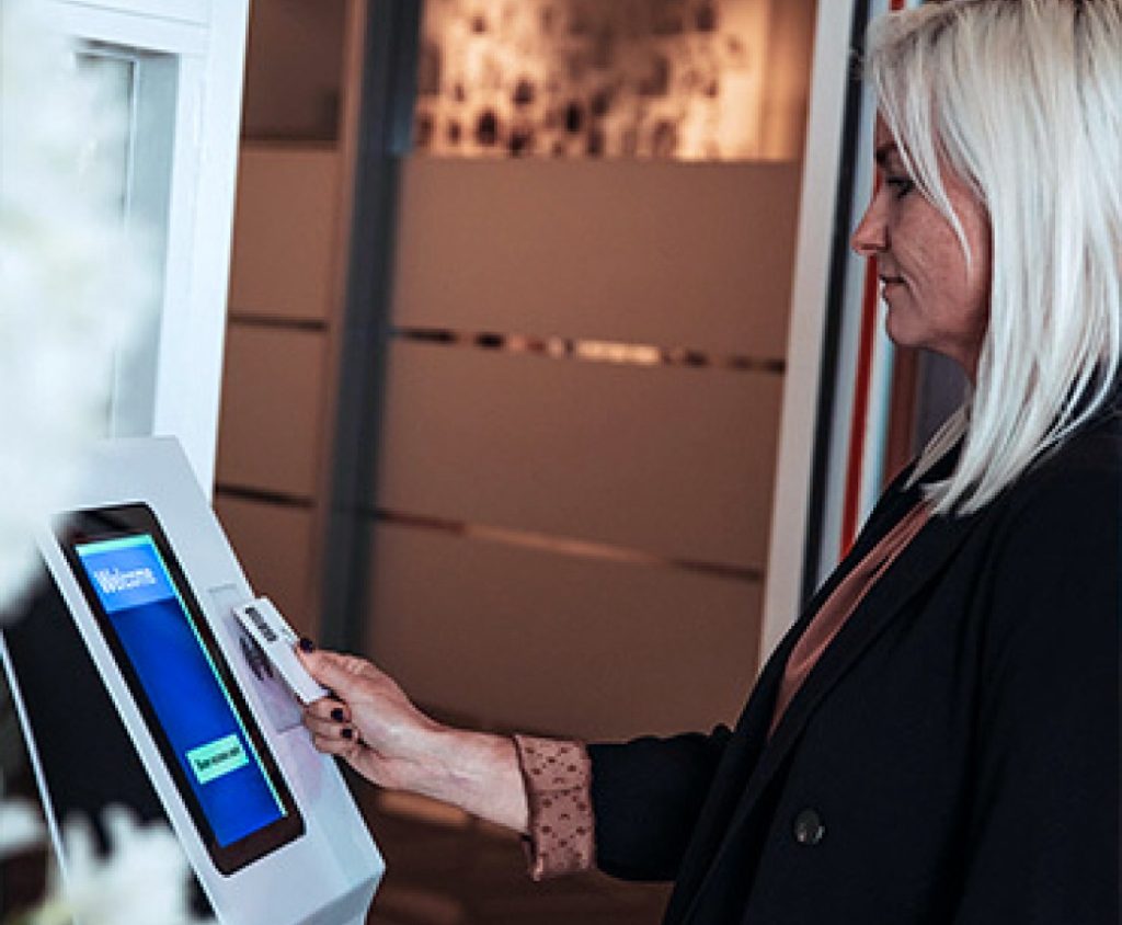 A woman with blonde hair uses a card to access a touchscreen kiosk.