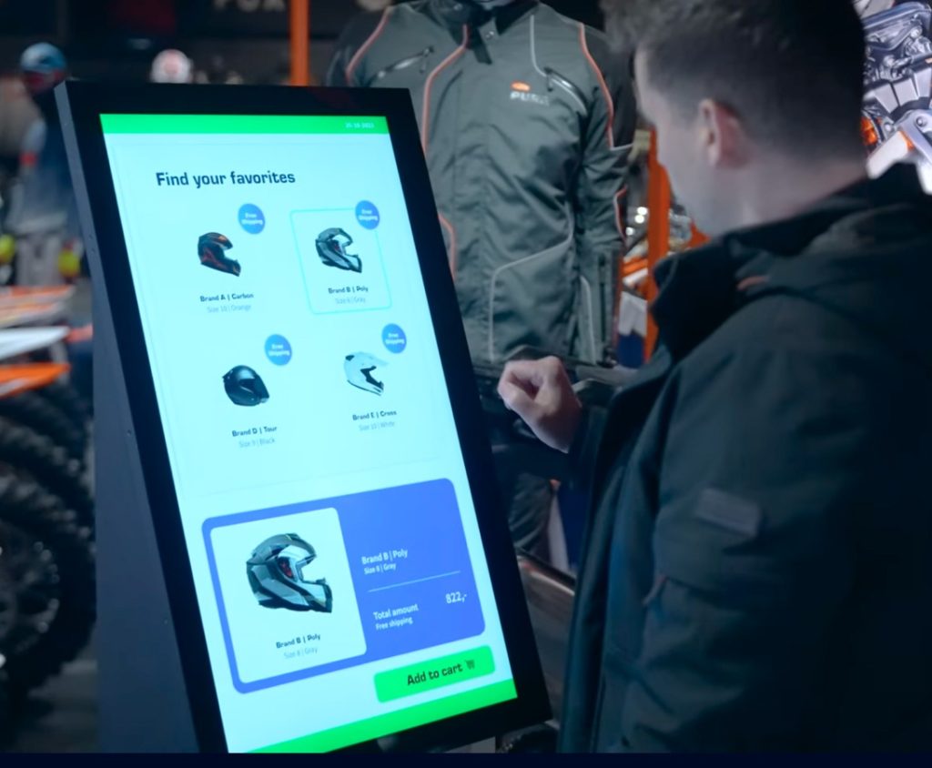 A person interacts with a touchscreen display in a store, selecting a helmet from a list of options including Brand A, Brand B, and Brand C. The screen shows helmet details and has an "Add to cart" button.