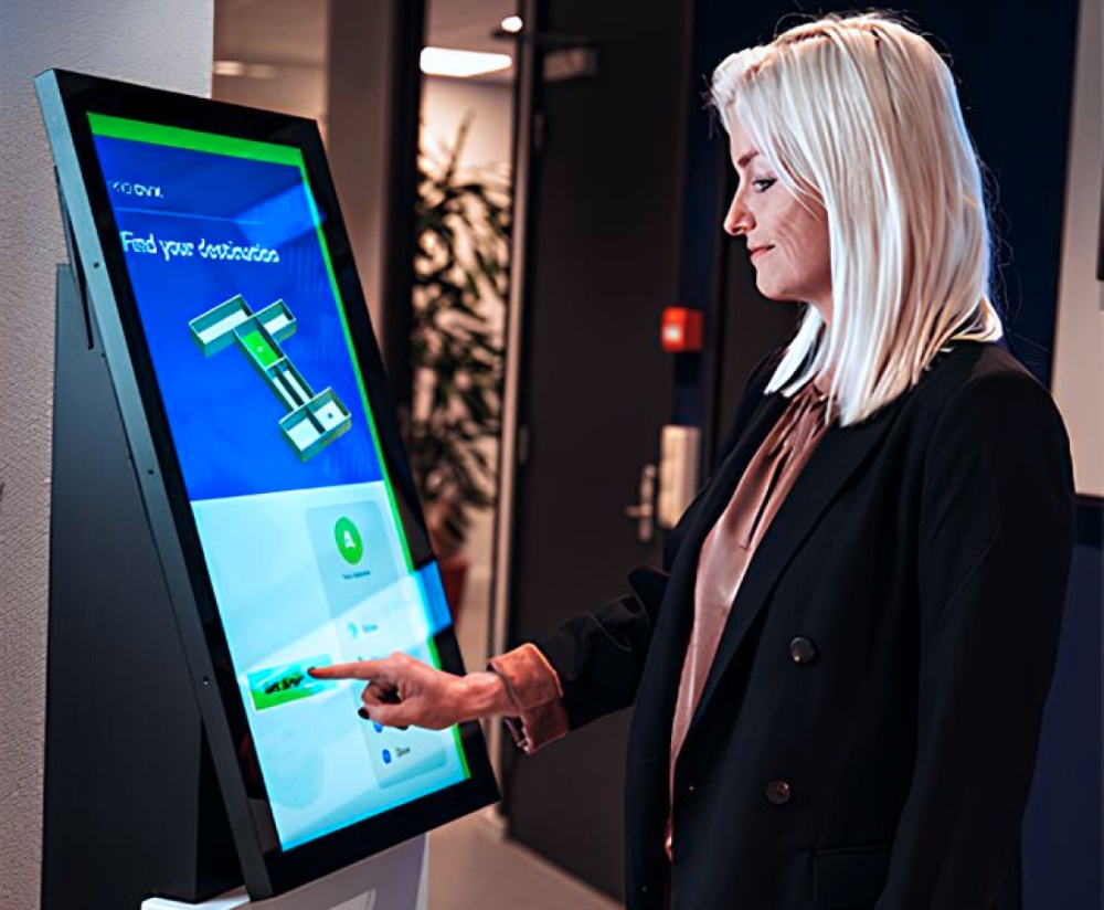 A woman with blonde hair uses a touchscreen kiosk in an indoor setting.