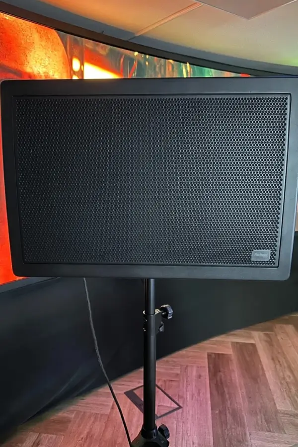A black rectangular speaker on a stand, positioned indoors with a wood-patterned floor and colorful background lighting.