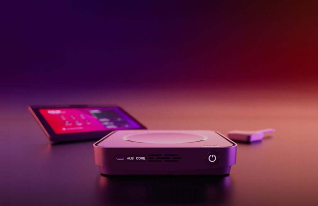 A compact electronic device labeled "HUB CORE" sits on a surface, with a tablet displaying a colorful interface and a small dongle in the blurred background.