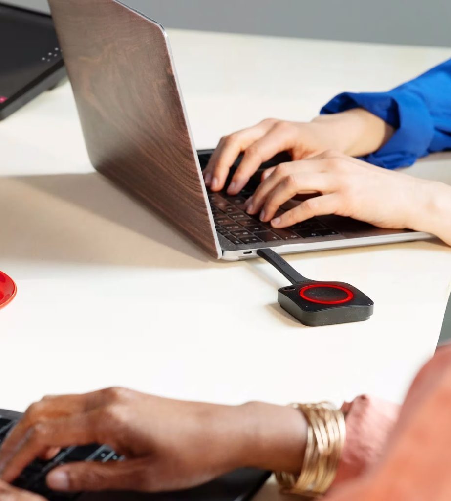 Two people are working on laptops at a desk; a wireless presentation device with a red circle is plugged into one of the laptops.