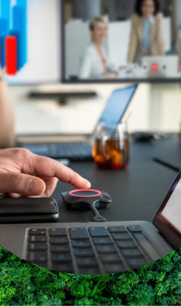 A hand presses a round button on a small device next to a laptop keyboard; two laptops, a glass mug, and a screen showing people are visible in the background.