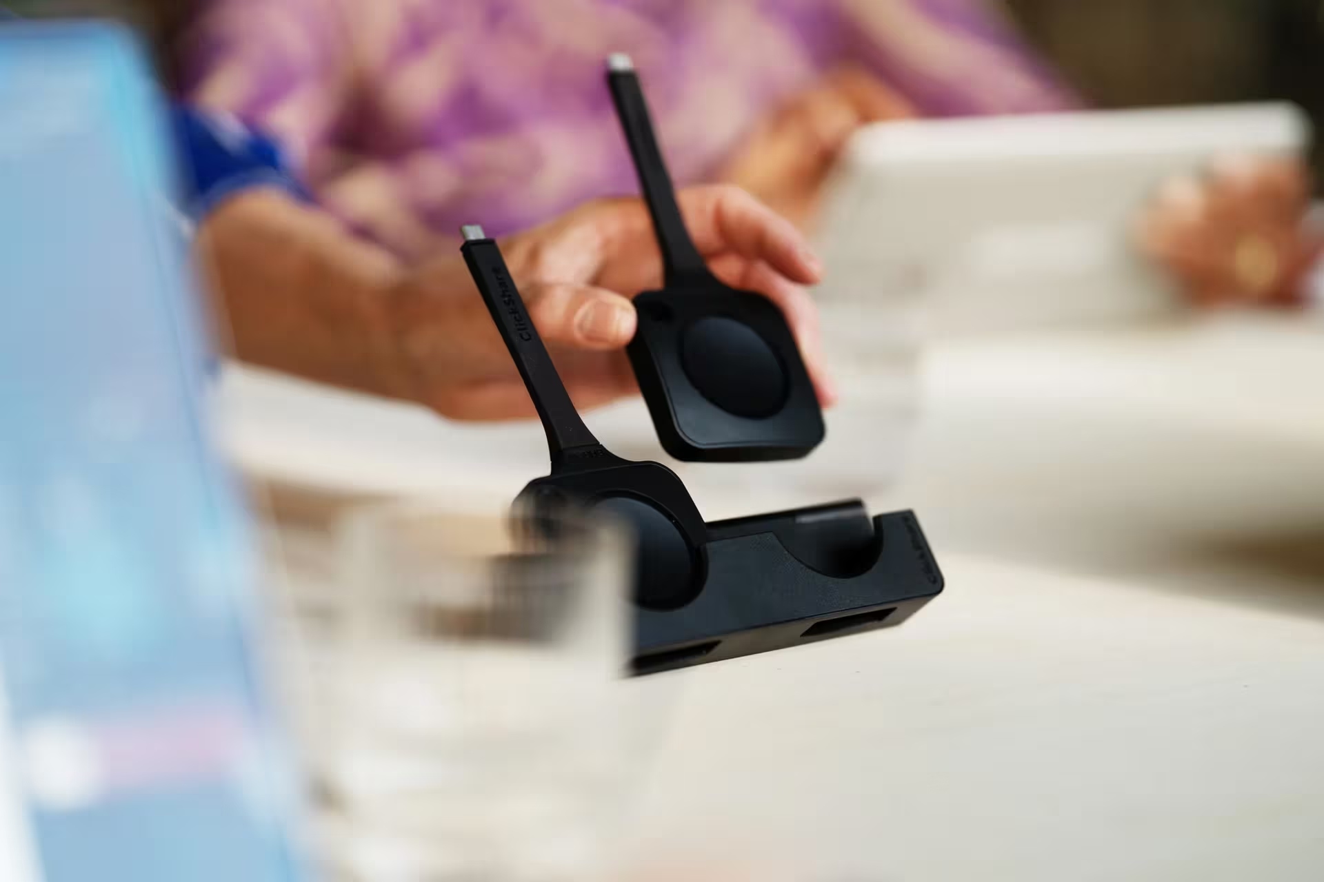A hand picks up a wireless presentation device from a black charging dock on a table, with blurred devices and glasses in the background.