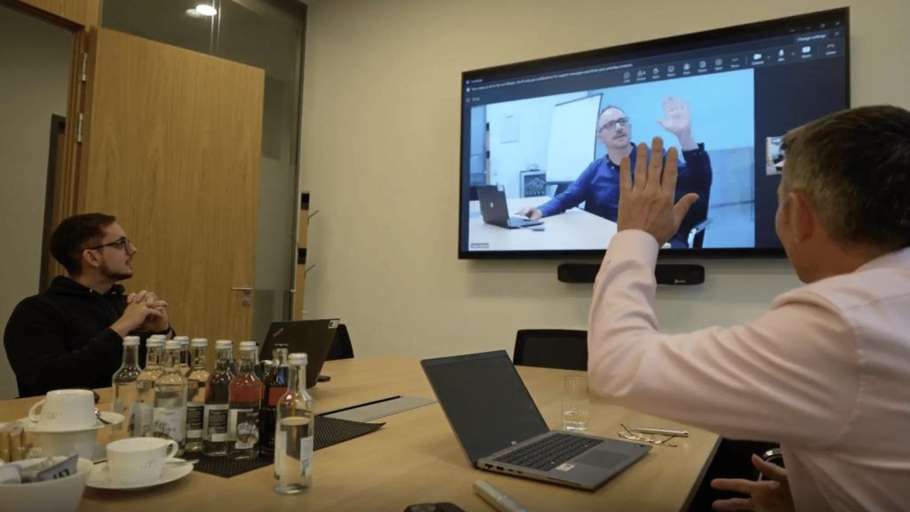 Two ifm staff in a meeting room with a remote colleague on the wall display, using Barco ClickShare wireless conferencing