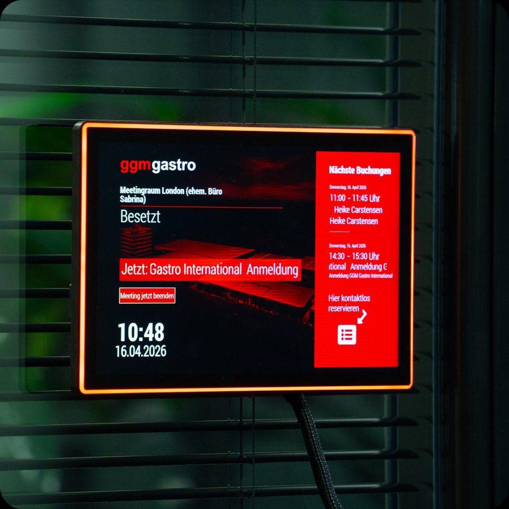 ProDVX APPC-10SFN touch panel mounted outside a GGM Gastro meeting room, LED bar lit red to show the room is occupied, with the next bookings listed on screen