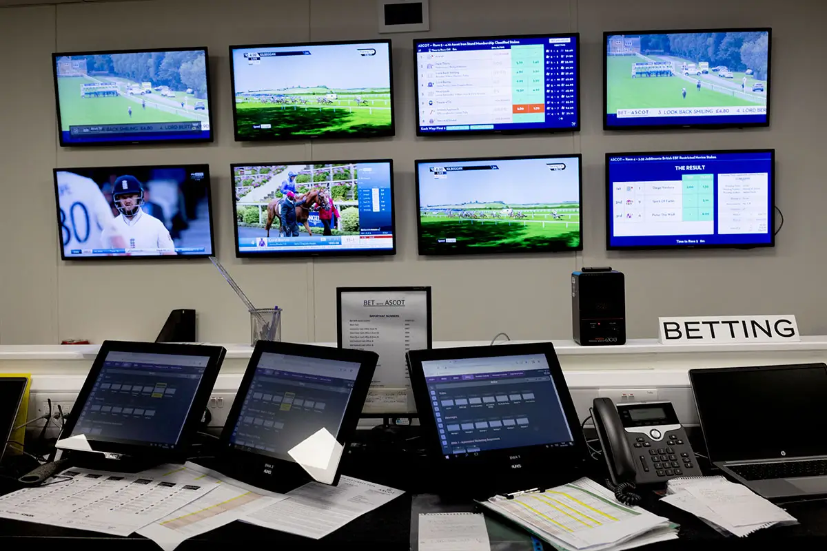 A control room with multiple screens displaying horse races and sports, several monitors on desks, papers, telephones, and a sign labeled "BETTING.
