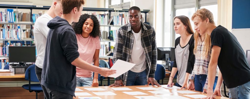 Six students stand around a table in a library, engaging in discussion and reviewing papers. Immersive AV spaces by The Avit Group and Marshall Cameras enhance the setting, with bookshelves and computers visible in the background.