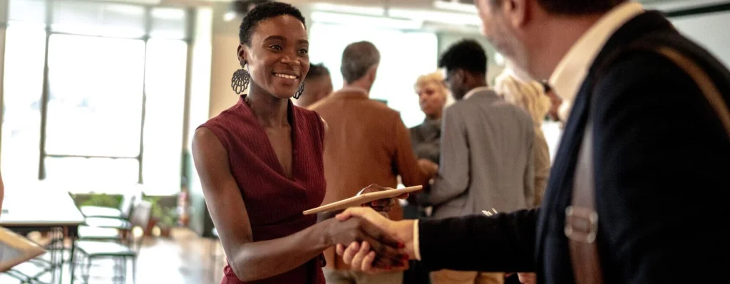 A woman smiles and shakes hands with a man while holding a plate at a Verizon networking event indoors, showcasing thoughtful event space management with several people in the background.