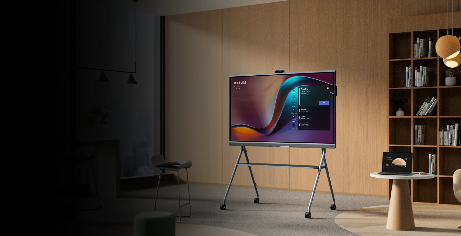 A large digital screen on a stand displays a user interface in a modern office with wooden walls, shelves, and minimal furniture.