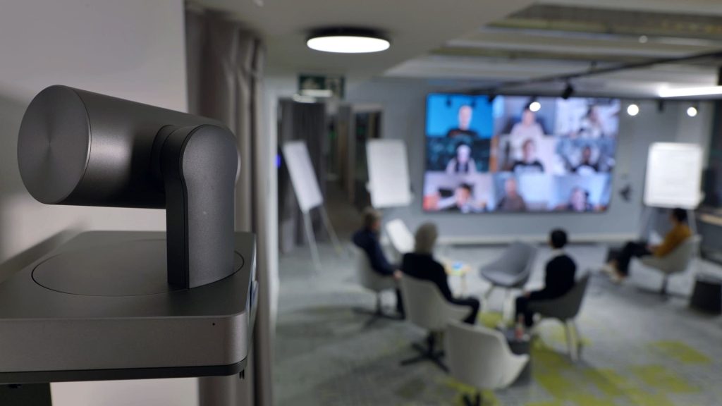 Yealink UVC PTZ camera in the foreground of a hybrid meeting room, with remote participants shown on a large display as staff sit in the room